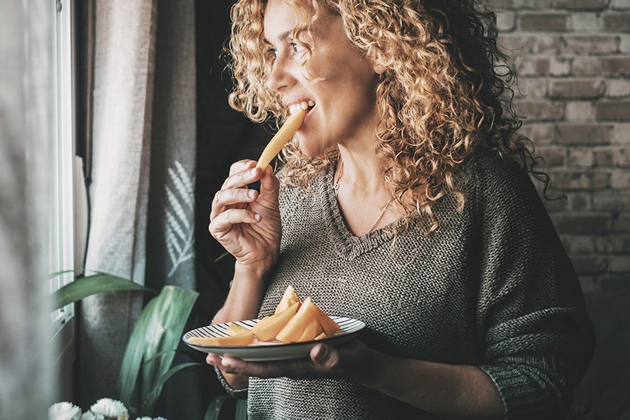 En kvinde st�r ved et vindue og spiser et stykke frugt.